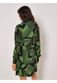 Grün und schwarz gemustertes Kleid mit geometrischem Design, langen Ärmeln, Kragen und Gürtel. Hergestellt aus glattem Stoff, mit figurbetontem Schnitt und geknöpften Manschetten.