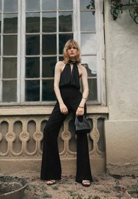 Zwart halter jumpsuit met een keyhole ontwerp, uitlopende benen, gladde stof, vergezeld van een kleine zwarte tas en metalen armbanden.