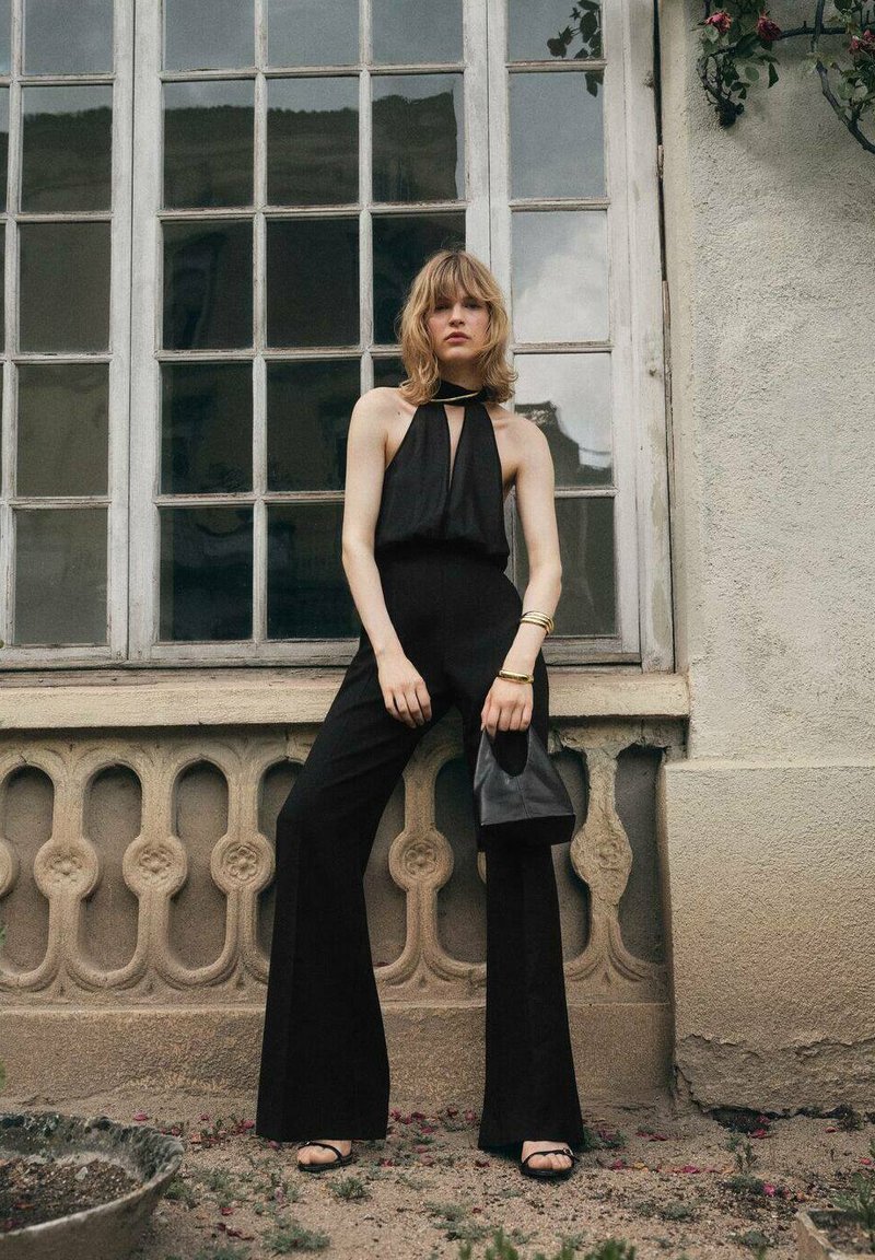 Zwart halter jumpsuit met een keyhole ontwerp, uitlopende benen, gladde stof, vergezeld van een kleine zwarte tas en metalen armbanden.