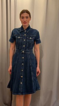 Woman standing in front of beige curtains wearing a button-up dark blue denim dress with short sleeves and chest pockets.