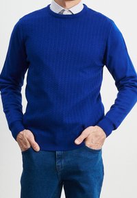 Sininen neulepusero, jossa on teksturoitu kuvio, pyöreä pääntie ja pitkät hihat. Yhdistetty sinisiin farkkuihin, joissa on ribbineuloskalvosimet ja -helma.