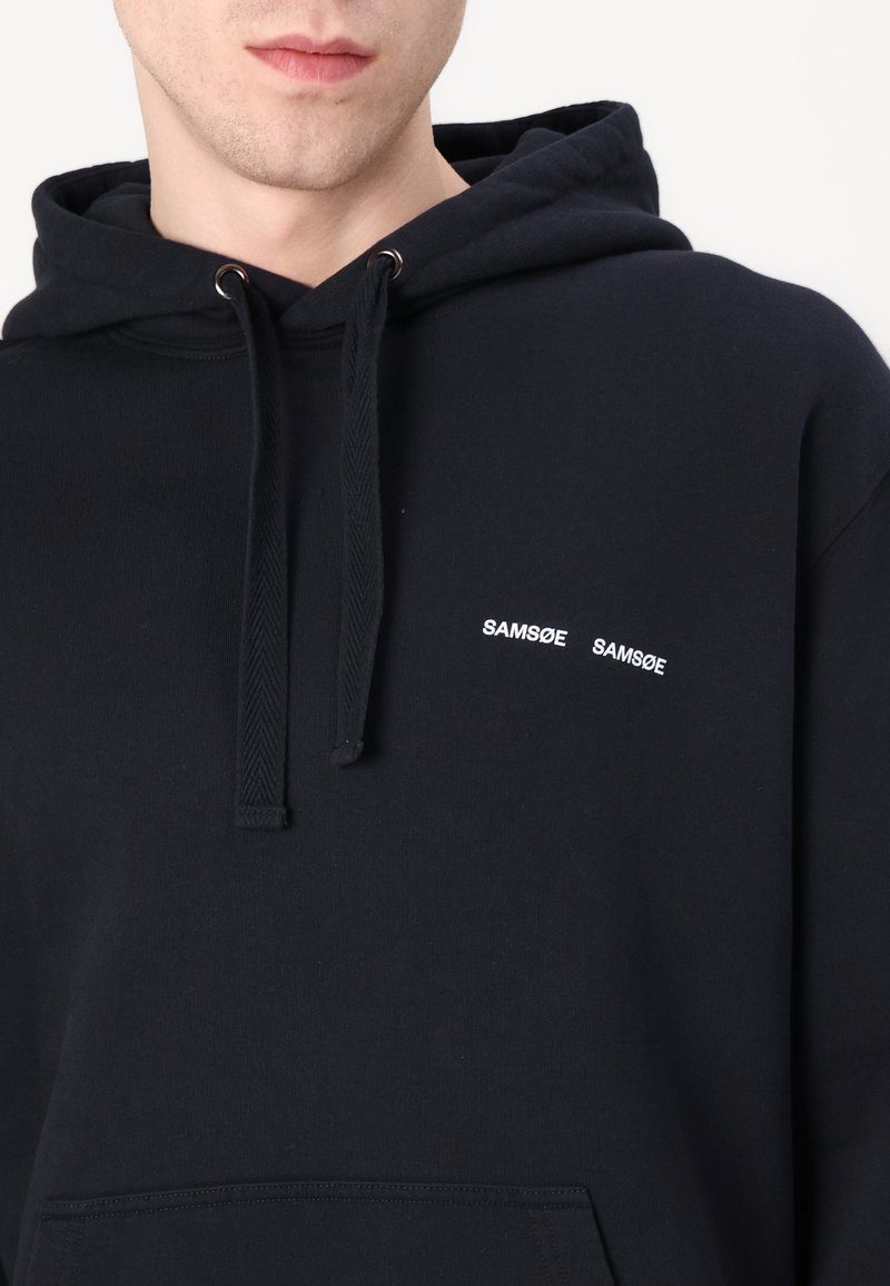Granatowy hoodie wykonany z miękkiej tkaniny z kieszenią typu kangur. Posiada białe haftowane napisy "SAMSOE SAMSOE" na klatce piersiowej oraz kaptur ze sznurkiem.
