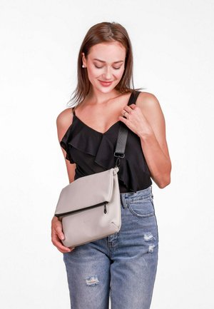 Šedá kožená taška přes rameno s překlopným víkem a černým popruhem. Modelka má na sobě černý volánový top a modré opotřebované džíny.
