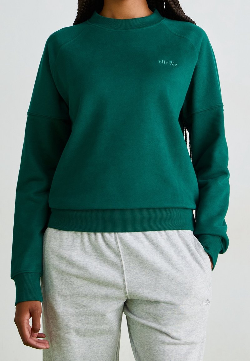 Personne portant un sweat-shirt col rond vert foncé et un pantalon de jogging gris clair, debout avec une main dans la poche devant un fond uni.