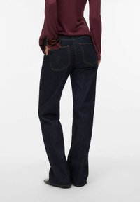 Mörk denimjeans med rak bendesign, som har två bakfickor och kontrasterande gula sömmar. Bärs med en långärmad topp.