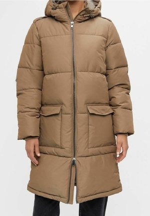 Long manteau d'hiver matelassé marron avec capuche, fermeture éclair frontale, poches à rabat, porté par une personne debout devant un fond uni.