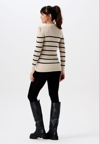 Beige gerippter Pullover mit schwarzen Streifen, taillierter Schnitt, lange Ärmel und runder Ausschnitt, kombiniert mit schwarzen Leggings und knielangen schwarzen Stiefeln.