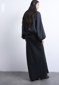 Γυναίκα με μακριά σκούρα μαλλιά, φορώντας μπλε μπλούζα με ρίγες και φουσκωτά μανίκια και μαύρη μακριά φούστα, όρθια μπροστά σε έναν λευκό τοίχο.