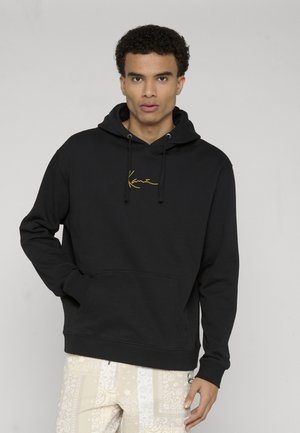 Jeune homme aux cheveux bouclés portant un sweat à capuche noir avec un logo script jaune et un pantalon crème à motifs, debout devant un fond uni.