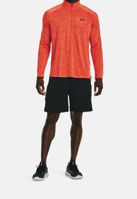Man wearing orange quarter-zip long sleeve shirt, black athletic shorts, and black running shoes standing on white background.