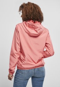 Giacca leggera con cappuccio di colore corallo, dotata di polsini e orlo elasticizzati, tessuto morbido e design casual senza motivi visibili.