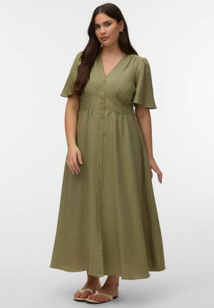 Ženska z dolgimi temnimi lasmi, oblečena v olivno zeleno maksi obleko z gumbi in kratkimi rokavi ter bež sandale, na enobarvnem ozadju.