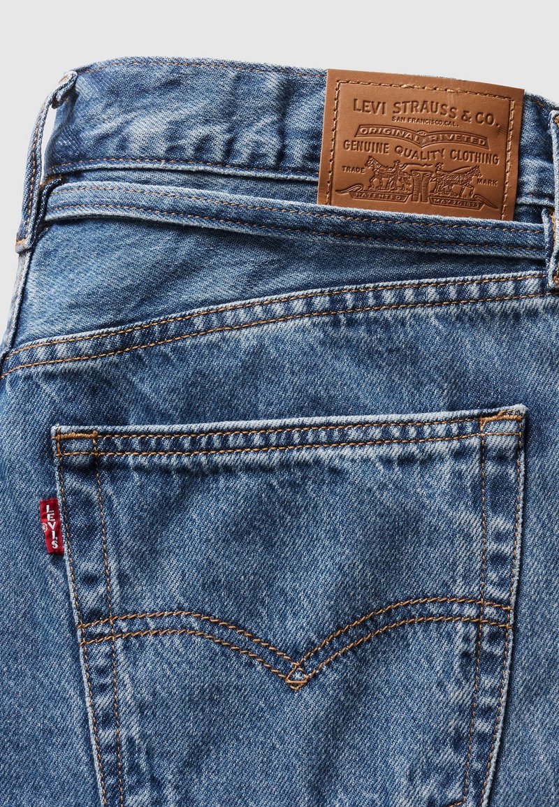 Jeans en denim de couleur bleu clair, design classique à cinq poches, couture orange et une étiquette en cuir marron avec les détails de la marque au niveau de la taille.