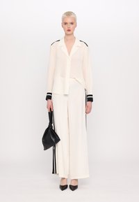 Bluză din material ușor și transparent, de culoare crem, cu accente negre, asociată cu pantaloni crem din material larg și pantofi cu vârf ascuțit negri. O geantă neagră ținută în una dintre mâini.