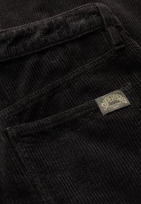 Zwart corduroy stof met gestructureerde ribbels, met een zichtbare zak en een genaaide label met merknaam en details.