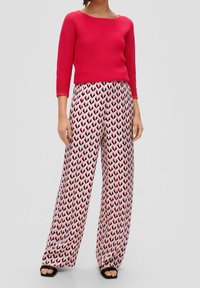 Donna che indossa un top a coste rosa brillante con maniche a tre quarti e pantaloni larghi a fantasia nei colori rosa, nero e bianco, con sandali neri.