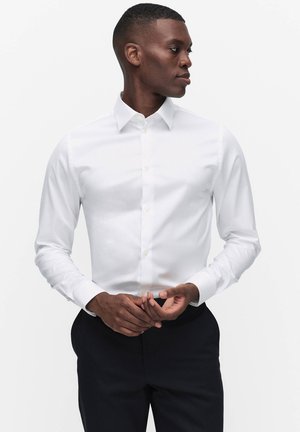 Mężczyzna ubrany w dopasowaną białą koszulę i czarne spodnie, patrzący na bok, poprawiający mankiety na tle jednolitego białego tła.