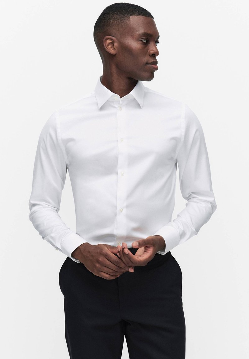 Mand iført en tilpasset hvid skjorte og sorte bukser, der ser sidelæns og justerer sine ærmer mod en ensfarvet hvid baggrund.