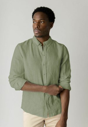 Mand med kort, krøllet hår iført lysegrøn, knappet skjorte, der ruller ærmerne op, ser til siden mod en ensfarvet lys baggrund.