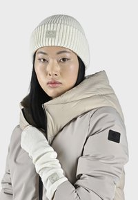 Doudoune beige, bonnet en tricot côtelé crème, gants sans doigts texturés et un logo noir prominent sur la manche de la doudoune.