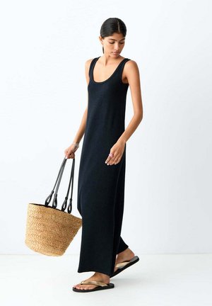 Frau in einem ärmellosen schwarzen Maxikleid, die eine große geflochtene Korbtasche hält, trägt Flip-Flops und geht vor einem einfarbigen weißen Hintergrund.