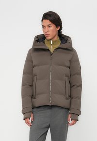 Brun polstret jakke med hætte, udstyret med lynlås og sidelommer. Båret over en grøn ribbet turtleneck og grå bukser.