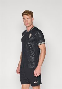 T-shirt de sport noir avec motif floral et design texturé, comprenant un col en V, des accents verts et un col rayé blanc. Associé à un short noir.