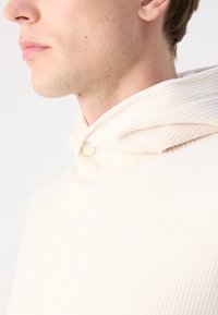 Prenda con capucha de color crema, con textura de canalé vertical, que presenta un cierre de botón en el cuello y una tela suave para mayor comodidad.