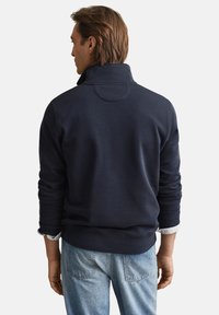 Sweatshirt azul-marinho com gola alta, punhos canelados e textura lisa. Apresenta um detalhe redondo nas costas, combinado com jeans claros.