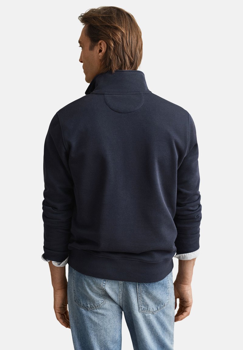 Marineblauer Pullover mit hohem Kragen, gerippten Bündchen und glatter Textur. Mit einem runden Akzent auf der Rückseite, kombiniert mit hellen Jeans.