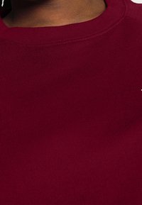 Bordeauxrode trui met ribbelstructuur, ronde halslijn en gestikte details. De stof lijkt zacht en licht elastisch. Geen opvallende logo's zichtbaar.