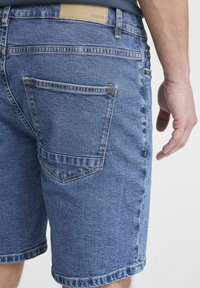 Pantalones cortos de mezclilla en azul medio, con un bolsillo trasero, detalle de costura y un parche de cuero con el nombre de la marca "ISOLID" en la cintura.