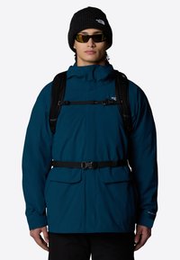 Chaqueta impermeable de color verde azulado con un cuello alto, cinturón ajustable y dos bolsillos frontales, llevada con un gorro negro y gafas de sol espejadas.