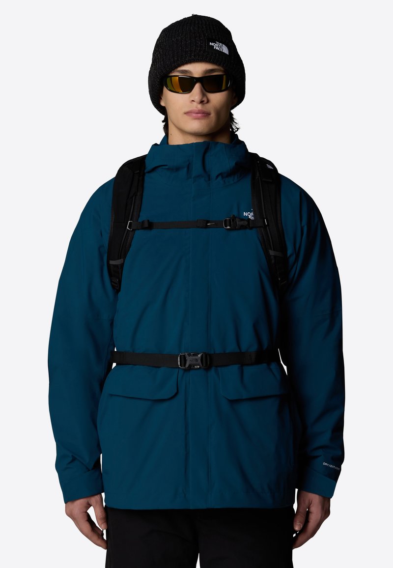 Chaqueta impermeable de color verde azulado con un cuello alto, cinturón ajustable y dos bolsillos frontales, llevada con un gorro negro y gafas de sol espejadas.