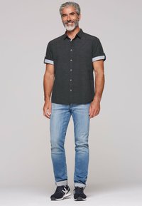 Korte mouwen, knoopshirt in donkergrijs met gestreepte manchetten, gecombineerd met lichtblauwe jeans en zwarte sneakers. Effen achtergrond.