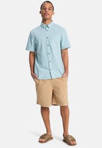 Ανοιχτό μπλε πουκάμισο με κοντά μανίκια και τσέπη στο στήθος, σε συνδυασμό με χακί σορτς και καφέ σανδάλια με λουράκια. Casual καλοκαιρινή εμφάνιση.