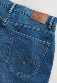 Zila džinsa biksšu aizmugurējā kabata ar brūnām šuvēm un brūnu ādas plāksnīti pie jostasvieta ar uzrakstu "DENIM SUPPLY".