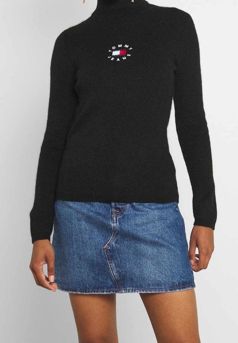 Czarny dziergany sweter z golfem z okrągłym logo „TOMMY JEANS”; zestawiony z jasnoniebieską mini spódnicą dżinsową z postrzępionym brzegiem.