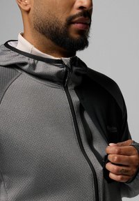 Szara kurtka w wzory z czarnym akcentem, zapinana na zamek z przodu, z kapturem. Posiada otwór na kciuk i subtelne logo marki na klatce piersiowej.