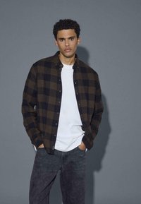 Bruine en zwarte geblokte overhemd met knopen over een wit T-shirt, gecombineerd met donkergrijze jeans. Glad materiaal met een casual pasvorm, voorzakken zichtbaar.