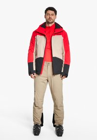 Skijacke in Rot, Beige und Schwarz mit hohem Kragen, Reißverschluss und kontrastierenden Ärmeln; kombiniert mit beigen Hosen und Skischuhen.