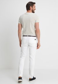 Pantalones de algodón blancos de corte slim, con costuras detalladas, dos bolsillos traseros y un cinturón marrón. Combinados con una camisa de manga corta gris claro.