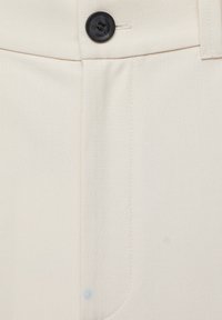 Close-up van de tailleband van beige broek met zwarte knoop, riemlus en zichtbare stikdetails.