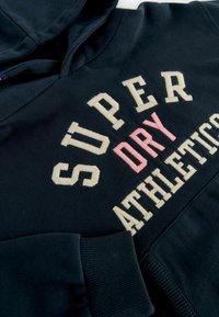 Námornícky modrá mikina s kapucňou vyrobená z bavlny, zdobená krémovo sfarbeným a ružovým výšivom s textom: "SUPER DRY ATHLETICS." Súčasťou sú rebrované manžety.