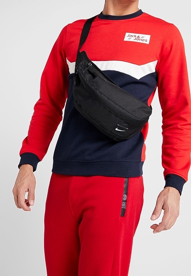 Sac en nylon noir porté en bandoulière, avec fermeture éclair, arborant un logo Nike et une sangle réglable, porté sur un sweat-shirt rouge et marine avec un pantalon assorti.