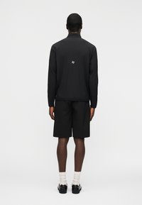 Άνδρας που στέκεται γυρισμένος ανάποδα, φορώντας μαύρο μακρυμάνικο μπουφάν με μικρό λογότυπο στο πίσω μέρος, μαύρο σορτς, λευκές κάλτσες και μαύρα παπούτσια.