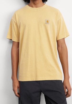 Mężczyzna ubrany w luźną, wyblakłą żółtą koszulkę Carhartt w połączeniu z czarnymi spodniami, stojący na tle jednolitego białego tła.