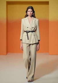 Beige linnen dubbelzijdig colbert met brede revers, ceintuur in de taille, gecombineerd met bijpassende wijde broek; model draagt zwarte hakken.