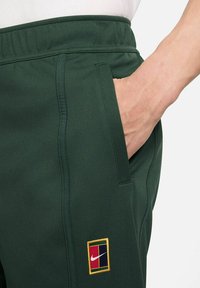 Pantalones de chándal verdes con cinturilla elástica, bolsillo lateral y una parche de logo de colores con acentos en rojo, azul y amarillo.
