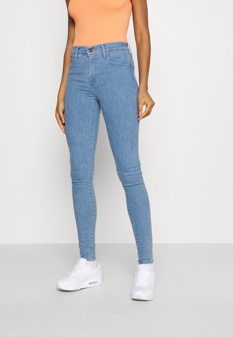 Personne portant un jean skinny bleu clair, des baskets blanches et un haut sans manches orange, debout devant un fond uni clair.
